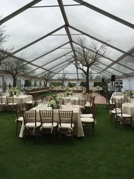 A clear-topped event tent set up over grass with arranged dining tables, chairs, and decorative bare trees.