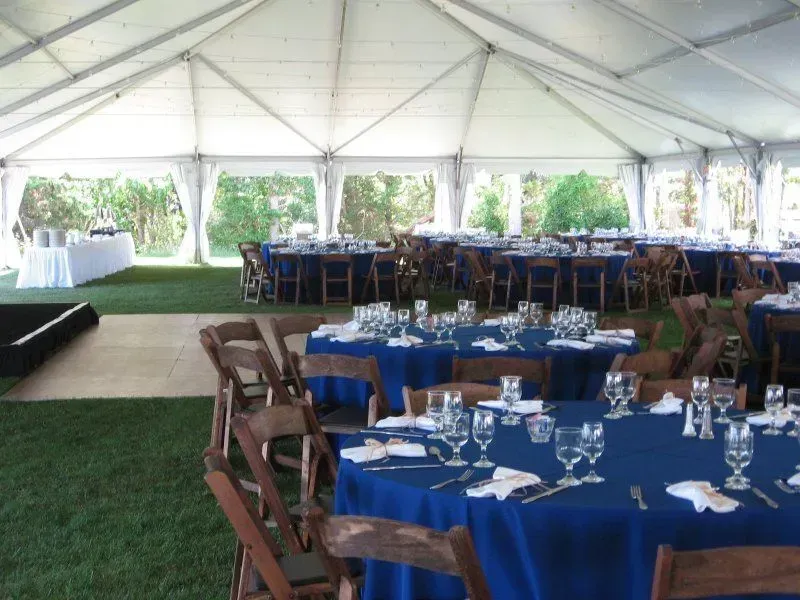 A large event tent set up with blue-clothed round tables and wooden folding chairs on a lawn for an outdoor gathering.