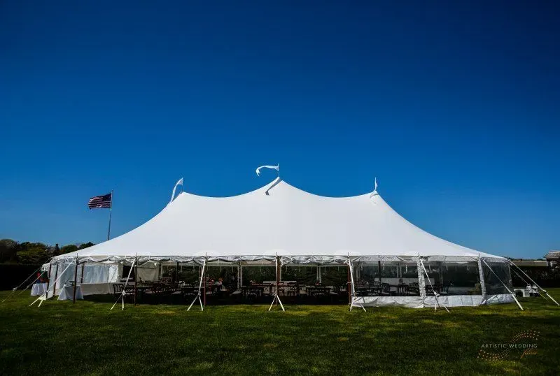 A large, white, multi-peaked event tent stands on a grassy lawn under a clear, bright blue sky.
