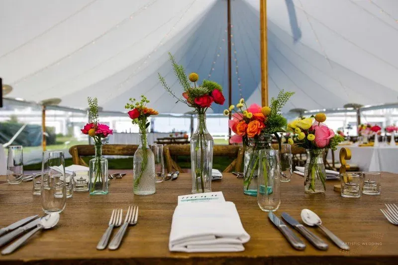 A rustic wooden table set for dinner under a tent, featuring floral arrangements in glass bottles and place settings.