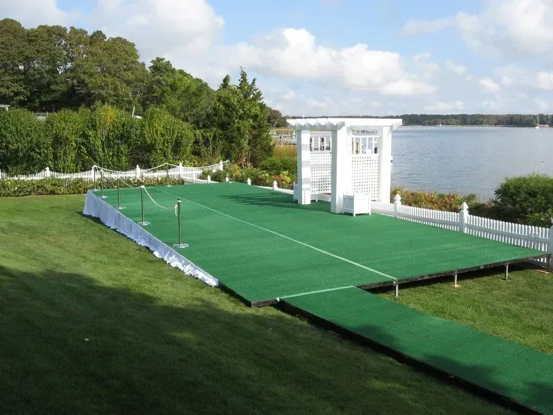 A raised green stage with a white wedding arch, set up on a grassy lawn overlooking a calm body of water.
