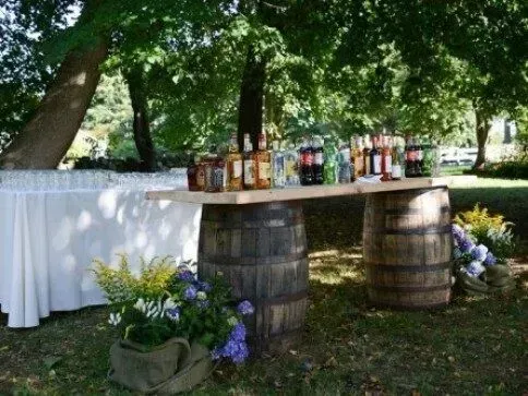 A bar setup outdoors with bottles of alcohol placed on a wooden plank resting across two wooden barrels.