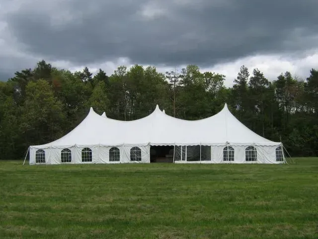 A large, white event tent set up on a green field in front of a line of trees under a cloudy, overcast sky.
