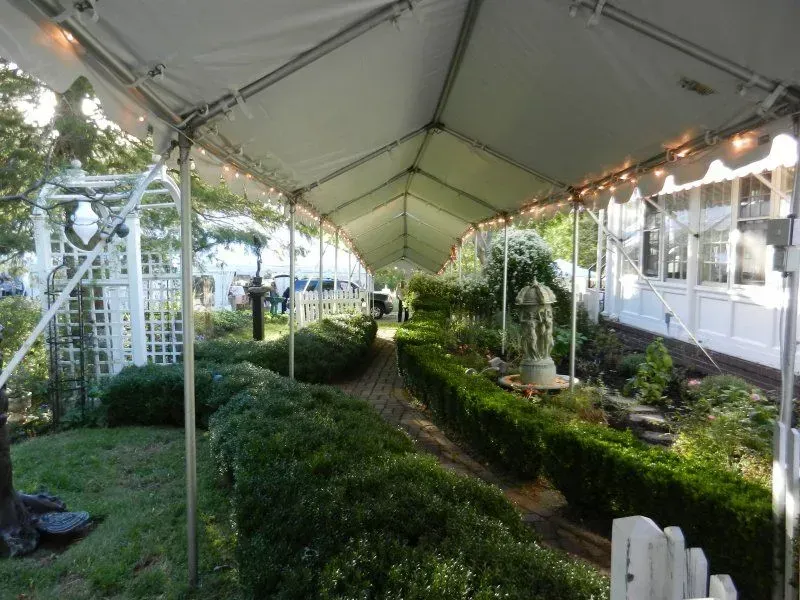 A pathway winds through manicured hedges under a long white event tent, leading to a garden statue beside a white building.