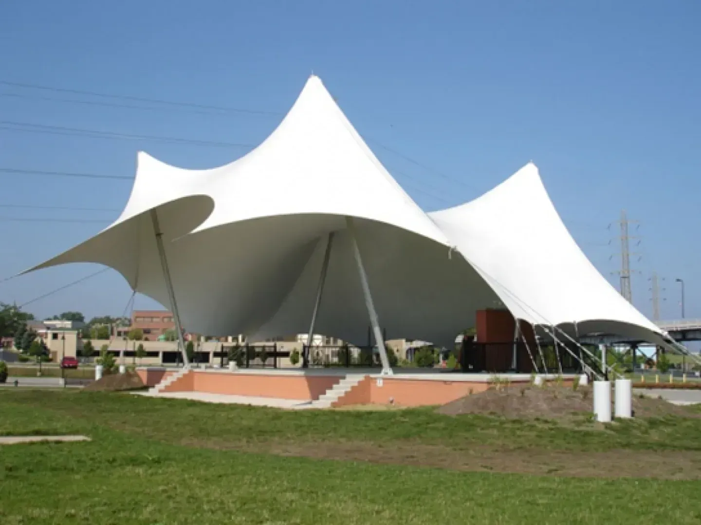 A white, multi-peaked tensile fabric amphitheater roof sits over a brick stage in a grassy outdoor park setting.