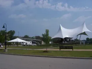 A park scene featuring a large white tensile fabric stage cover, a smaller event tent, and a bridge in the distance.