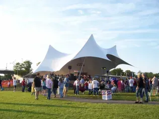 A crowd gathers on a lawn before a large, white, multi-peaked tensile stage canopy during an outdoor event.