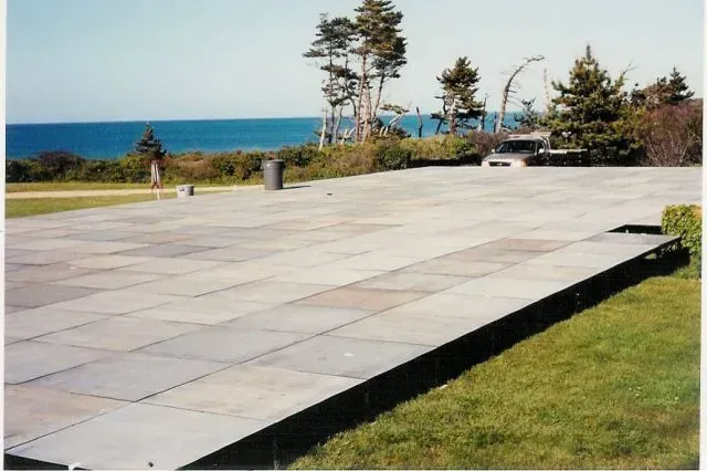 A wide, elevated stone patio overlooks a coastal landscape with green grass, trees, and the blue ocean in the distance.