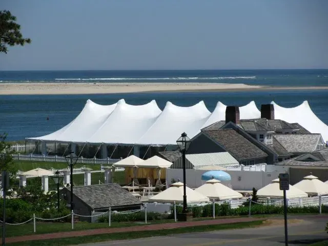 A large white event tent stands near the ocean, with several umbrellas and a coastal building visible in the foreground.