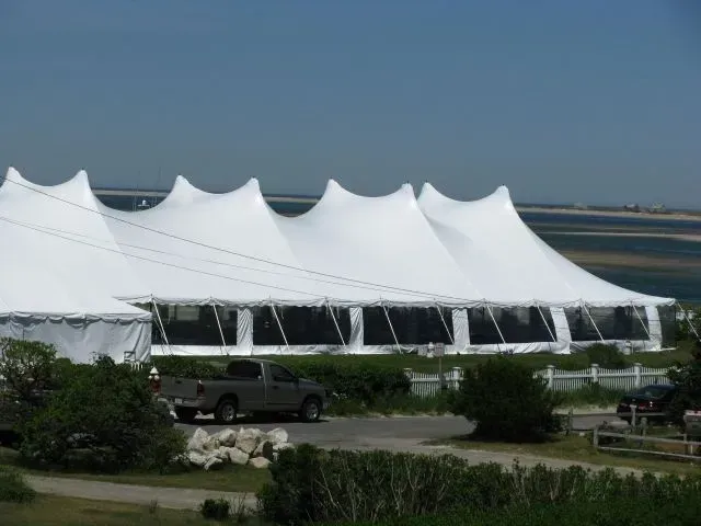 A large, white, multi-peaked event tent stands on a grassy area near a body of water, with a truck parked in front.
