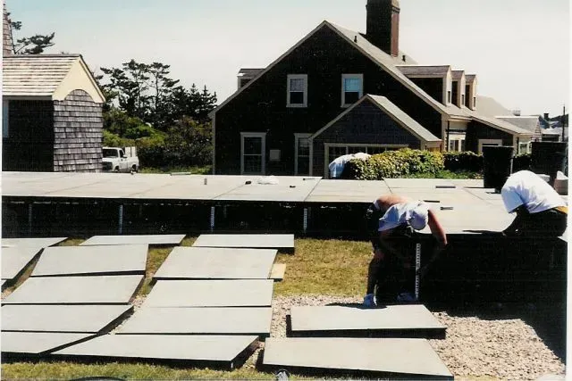 Two people set up a portable event stage on a lawn in front of a dark shingled house.