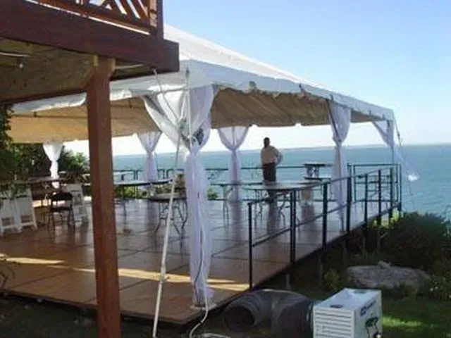 A white tent with draped fabric stands on a wooden deck overlooking the ocean, with a person standing by a railing.