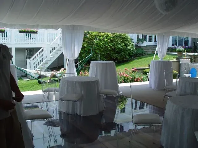 Outdoor event tent set up with white-clothed tables, chairs, and a hammock on a green lawn by a house with a deck.