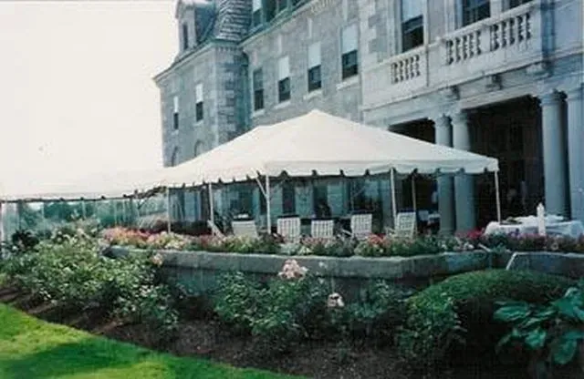 A large white event tent set up on a stone terrace in front of a historic stone building, surrounded by rose bushes.
