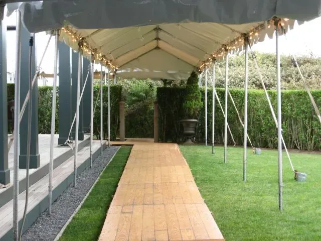 A long wooden path covered by a tent, flanked by a lawn and a grey gravel strip beside a house exterior.