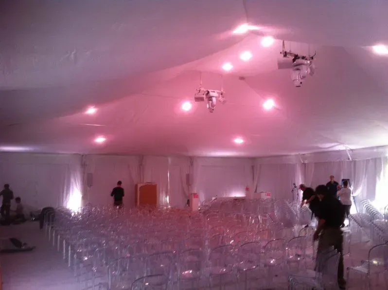 Event space with clear chairs set in rows under a white tent ceiling illuminated by pink lights and overhead projectors.