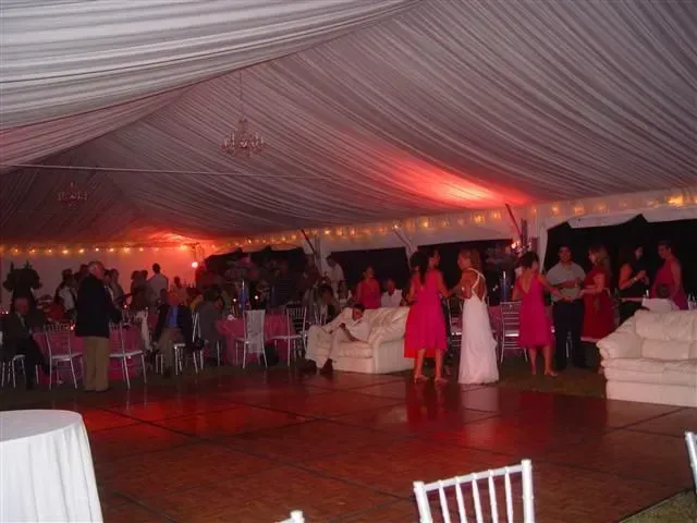 A wedding reception in a large white tent with guests dancing, socializing, and sitting on sofas near a wooden dance floor.