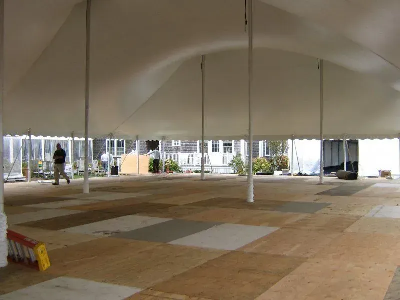 A large white tent with interior support poles and a wooden panel floor, set up outdoors on a lawn near a building.