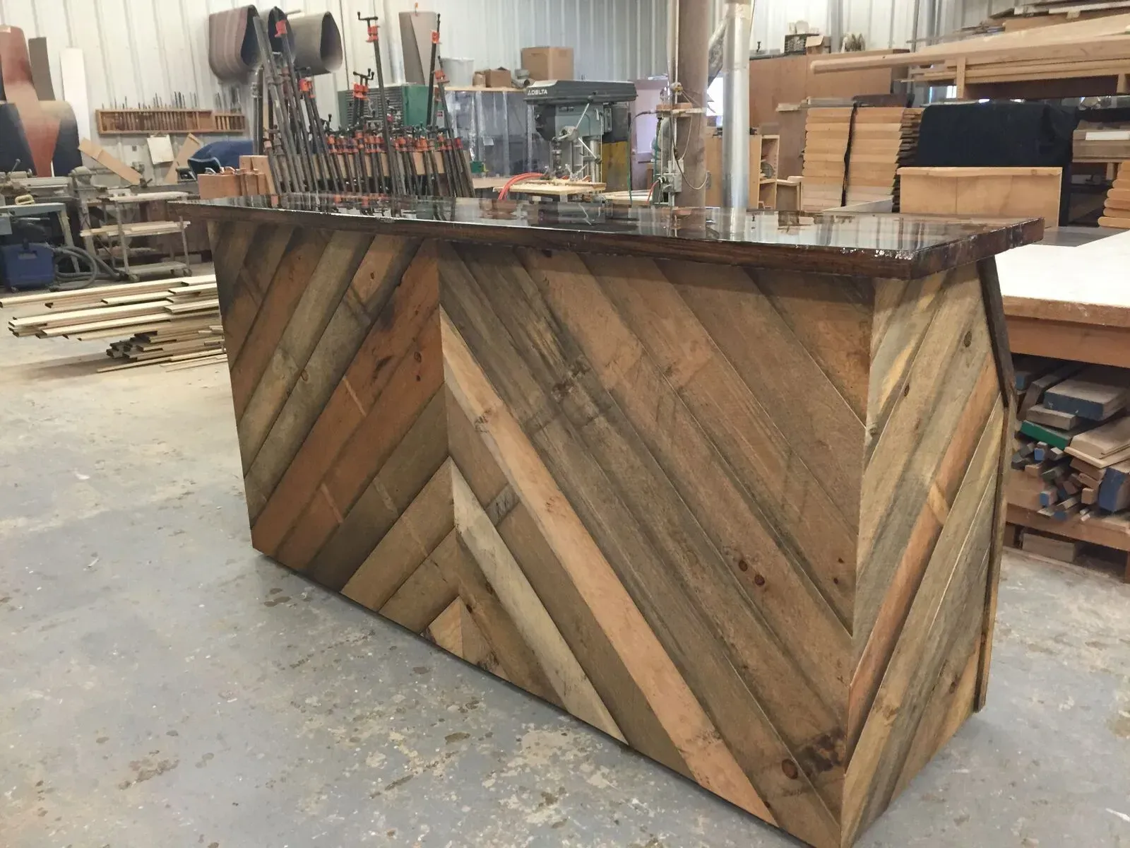 A custom wooden bar with a chevron pattern made of reclaimed planks, situated in a cluttered woodworking shop.