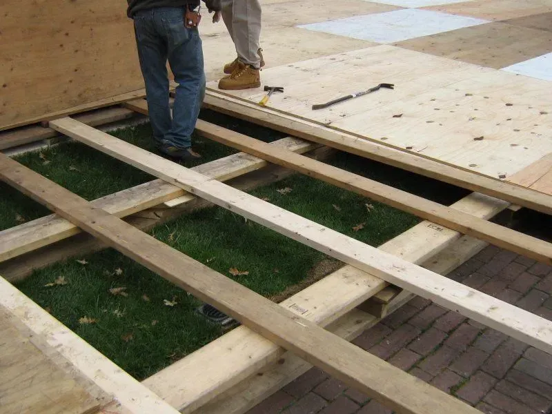 Two people stand on a partially constructed wooden floor platform installed over grass.