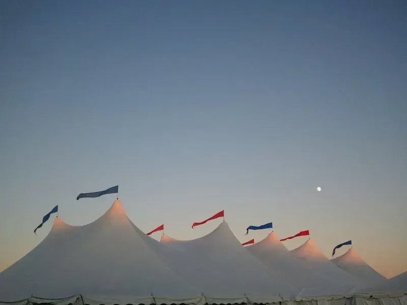 A large white event tent with multiple peaks topped by red and blue flags against a clear dusk sky with a visible moon.