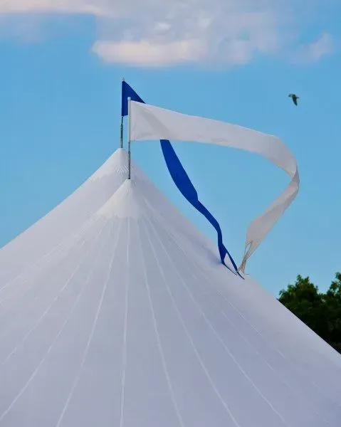 A white tent peak topped with a small blue flag and a long white streamer fluttering against a clear blue sky.