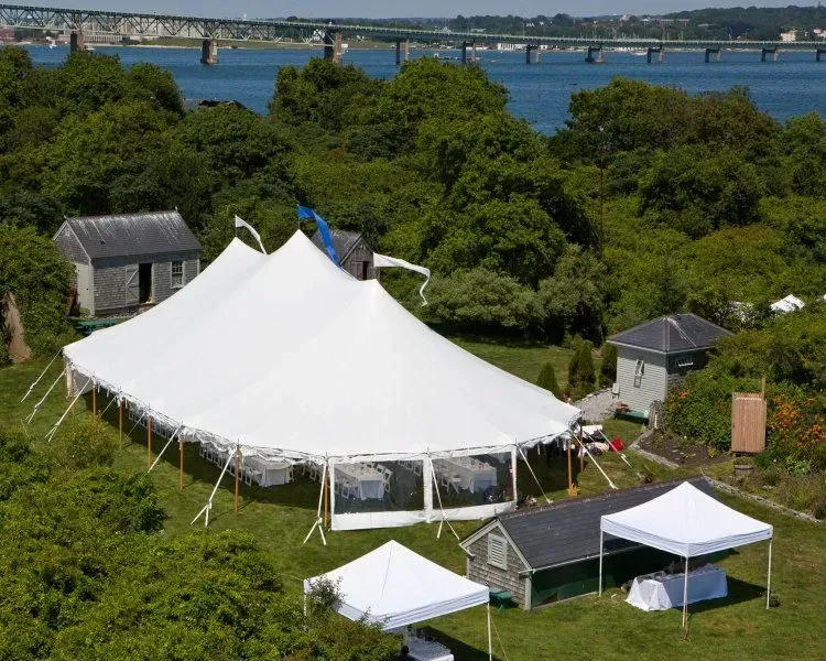 An aerial view of a large white party tent and two smaller pop-up tents set up on a grassy lawn near the waterfront.