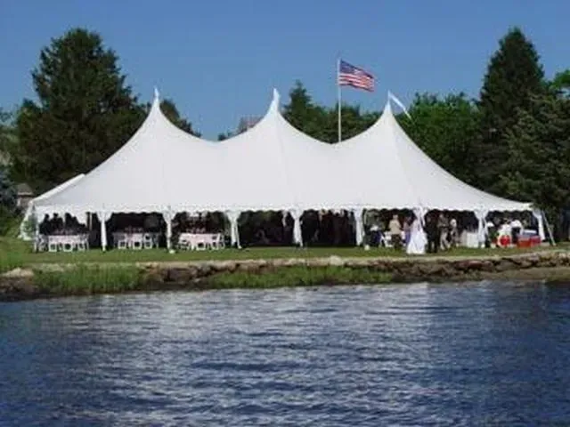 A large white event tent with three peak sections, set on a grassy riverbank under a blue sky with an American flag.