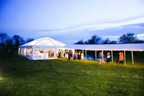 People gather under illuminated white event tents on a grassy field at dusk.