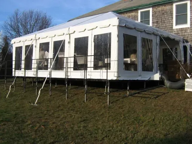 A large white event tent with transparent windows sits on an elevated metal platform next to a residential house.
