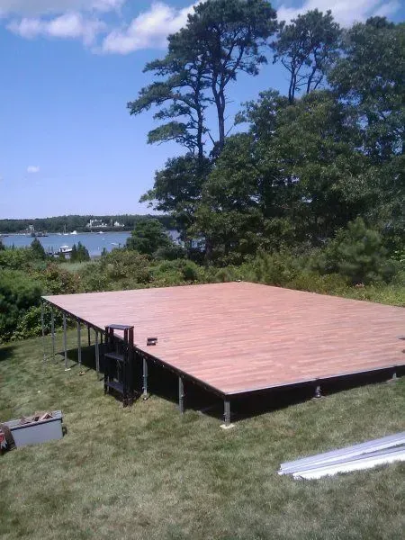 A large, elevated rectangular wooden platform sits on a grassy hill overlooking a body of water and distant trees.