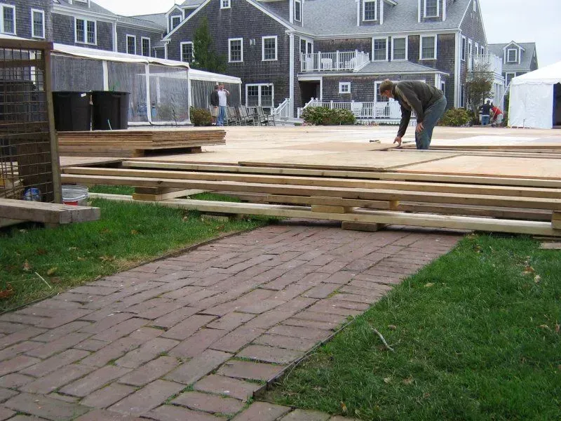 A worker kneels on a wooden subfloor construction project in a grassy lawn area with a brick path in the foreground.