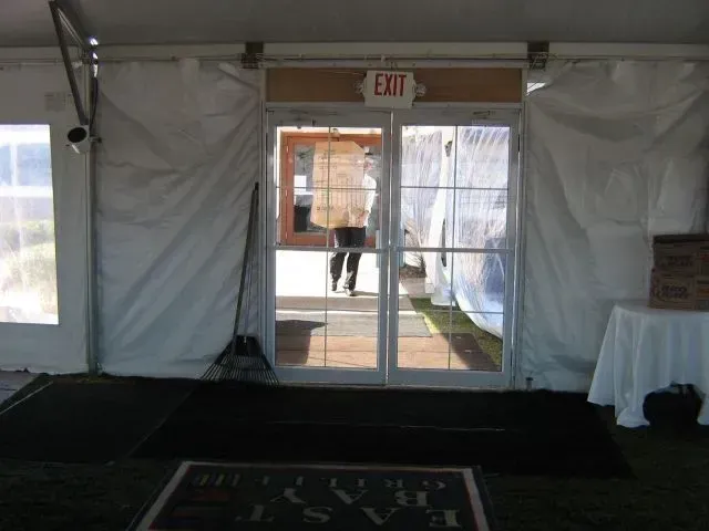 A person carries a large box through the open glass doors of a tented event space with a black floor mat.