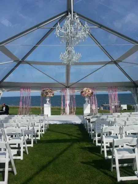 An outdoor wedding ceremony setup under a clear-roof tent, featuring white chairs, floral arrangements, and a chandelier.
