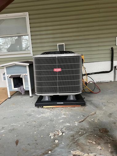 An outdoor air conditioning unit sits on a black mounting base against a house with light green horizontal siding.