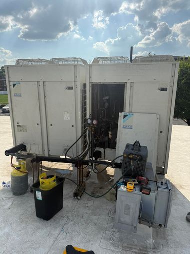 A technician works on an open Daikin HVAC unit on a rooftop, with refrigerant tanks and tools nearby.