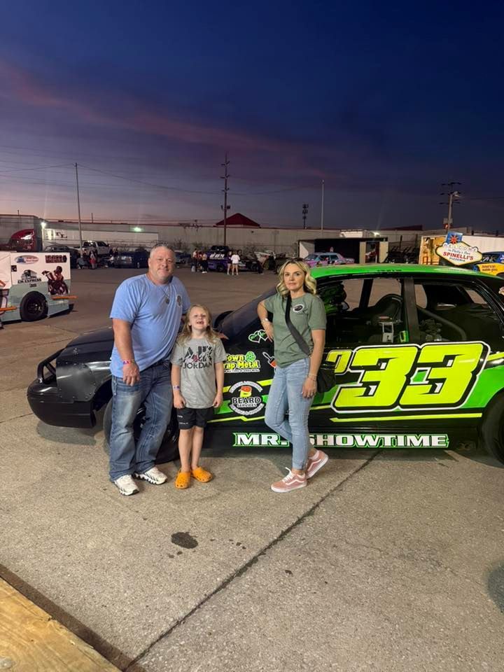 Three people standing together in a parking lot next to a black and neon-green race car labeled 