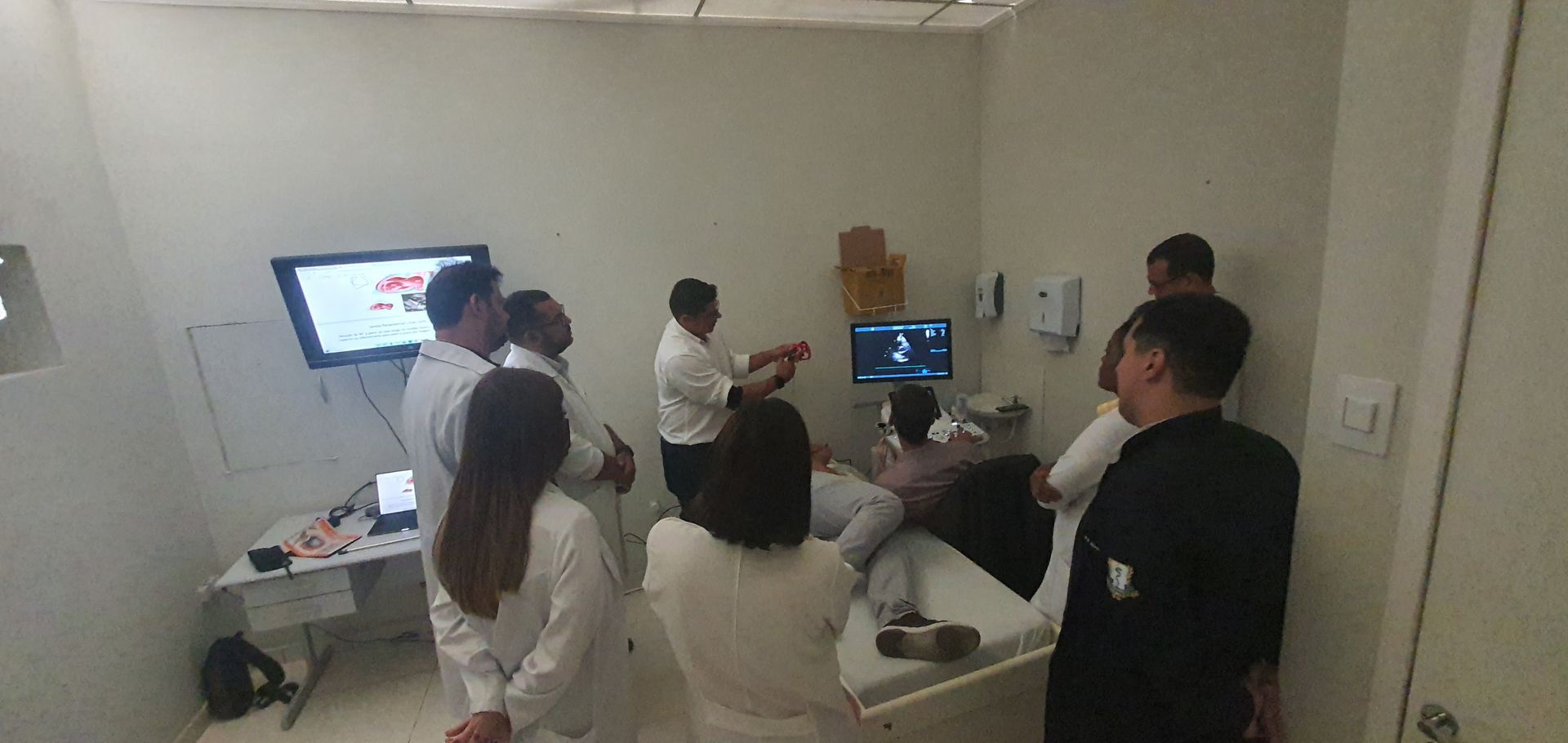 Sala de exames médicos com várias pessoas reunidas em torno de um paciente deitado em uma cama.