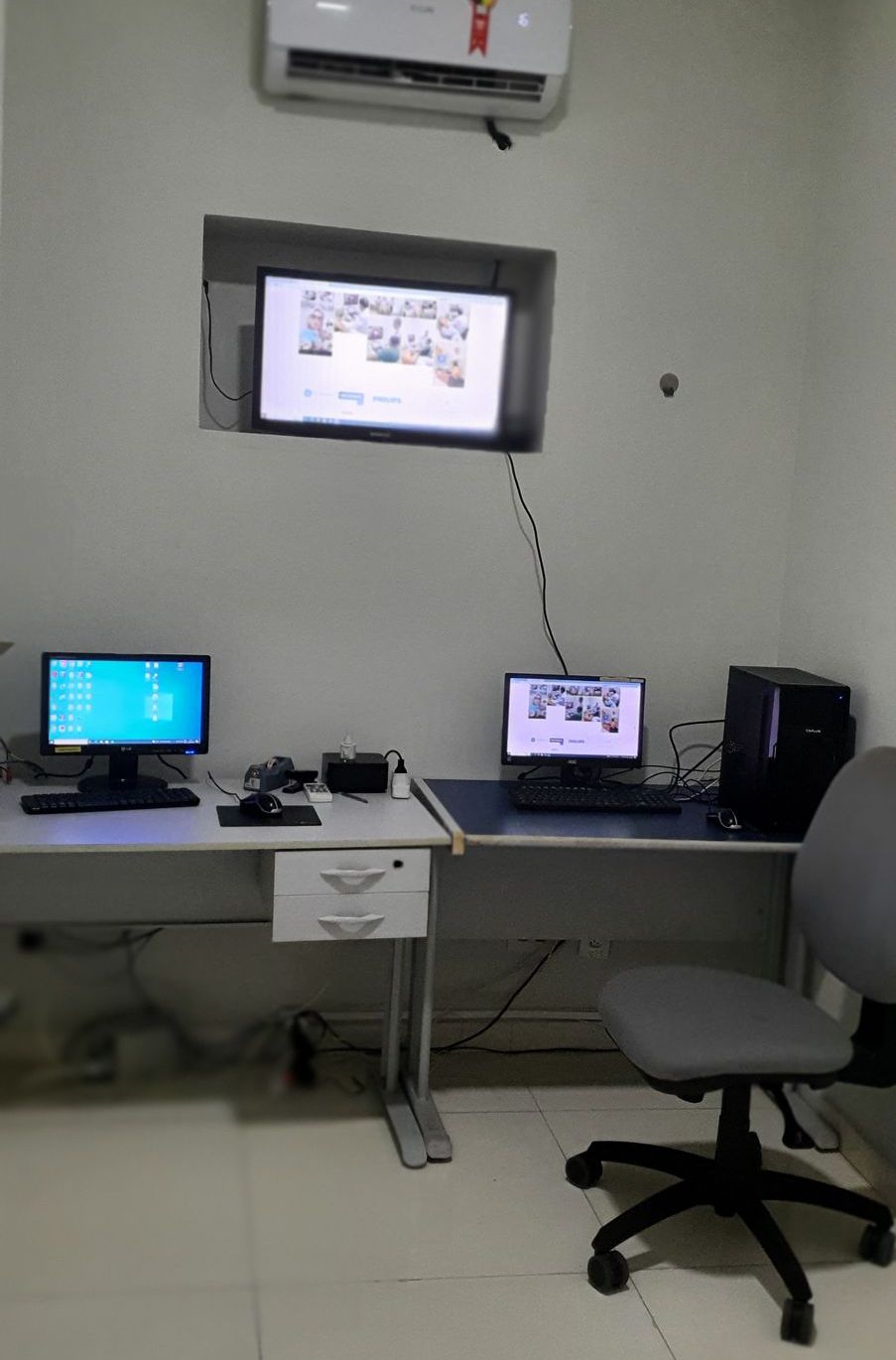 Sala com computadores, monitor na parede, mesa, cadeira e aparelho de ar condicionado.