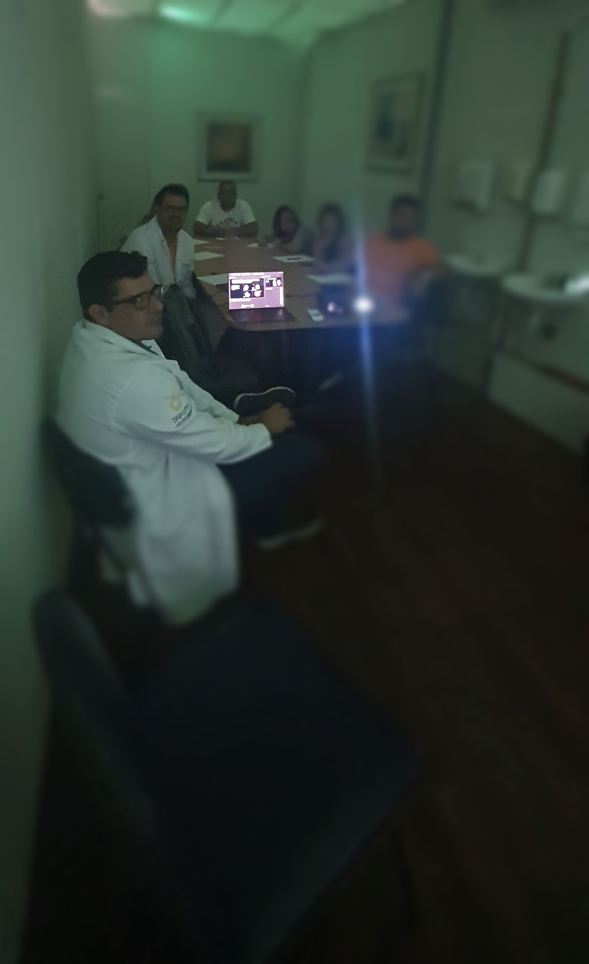 Pessoas de jaleco branco em uma mesa de conferência com um laptop, em uma sala com uma pia.