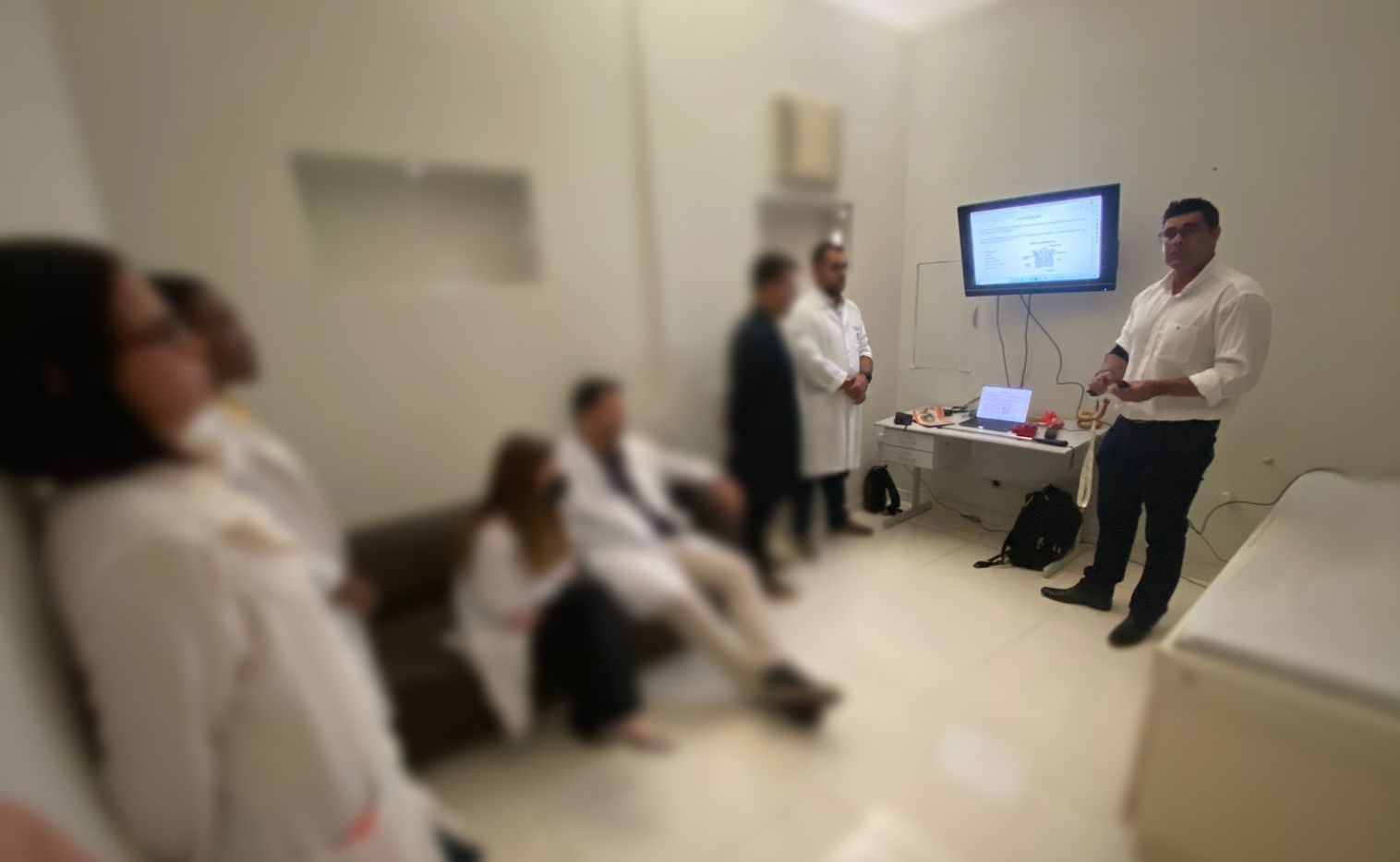 Homem fazendo uma apresentação para um grupo em uma sala com equipamentos médicos e uma tela exibindo dados.