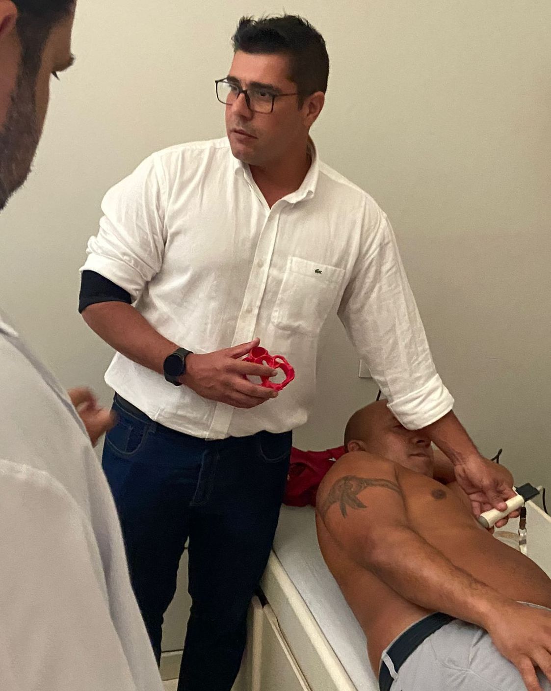 Homem de camisa branca examinando o tórax de outro homem em uma mesa de exame, com uma terceira pessoa presente.