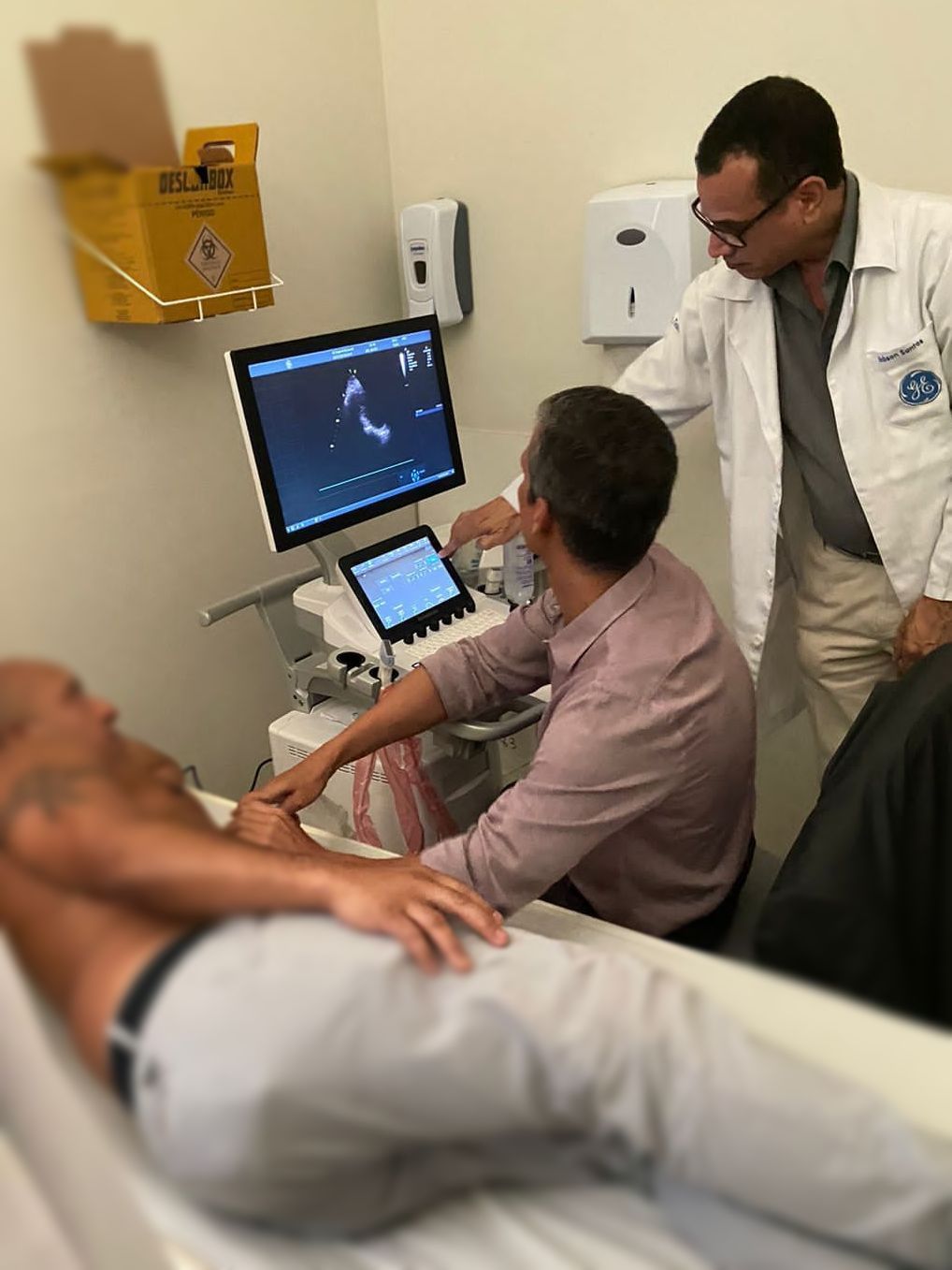 Dois médicos realizam um ultrassom em um paciente em uma sala de exames; um monitor exibe as imagens.