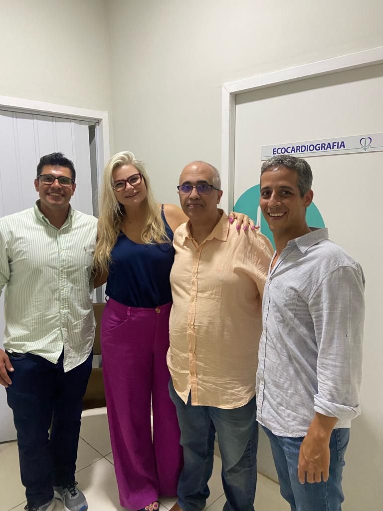 Quatro pessoas posam em um corredor. Dois homens nas laterais, dois no meio. A mulher veste calças rosa e uma blusa azul.