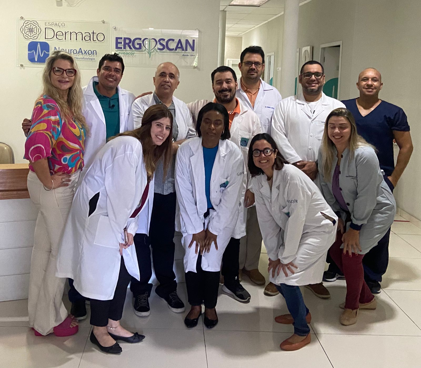 Grupo de pessoas de jaleco branco sorrindo em um escritório; placa com os dizeres