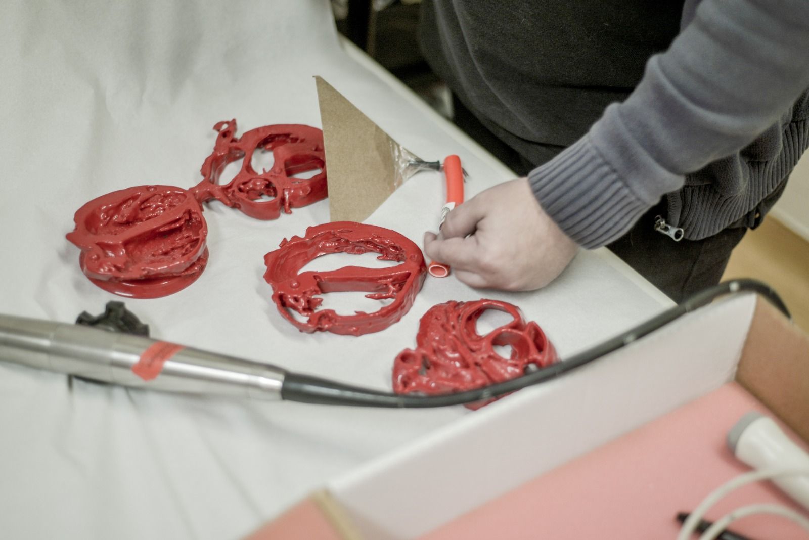 Pessoa montando peças de plástico vermelhas, impressas em 3D, sobre uma superfície branca, com ferramentas por perto.