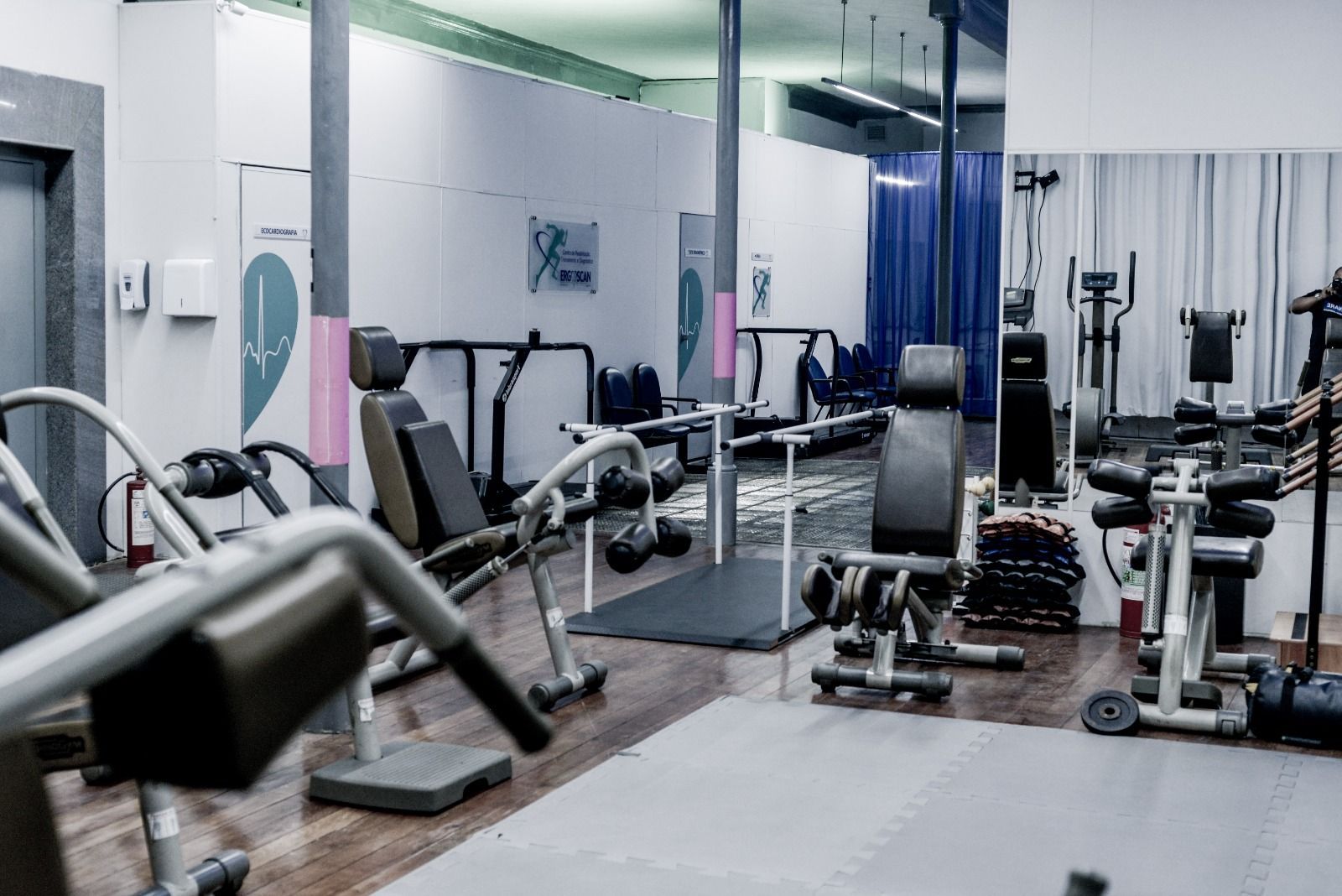 Interior de ginásio com equipamentos de exercício, incluindo pesos e máquinas, refletidos em um grande espelho.
