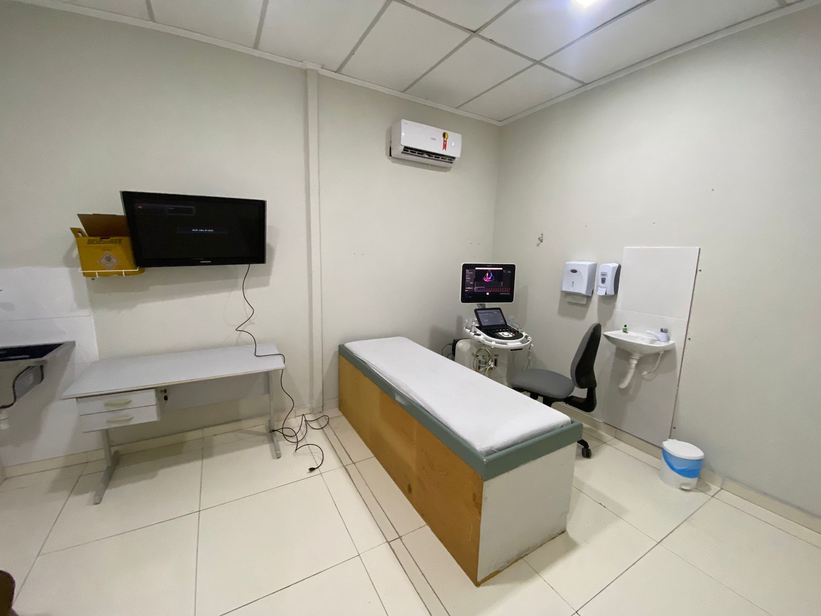 Sala de exame médico, aparelho de ultrassom, mesa de exame, monitor, pia e ar condicionado.