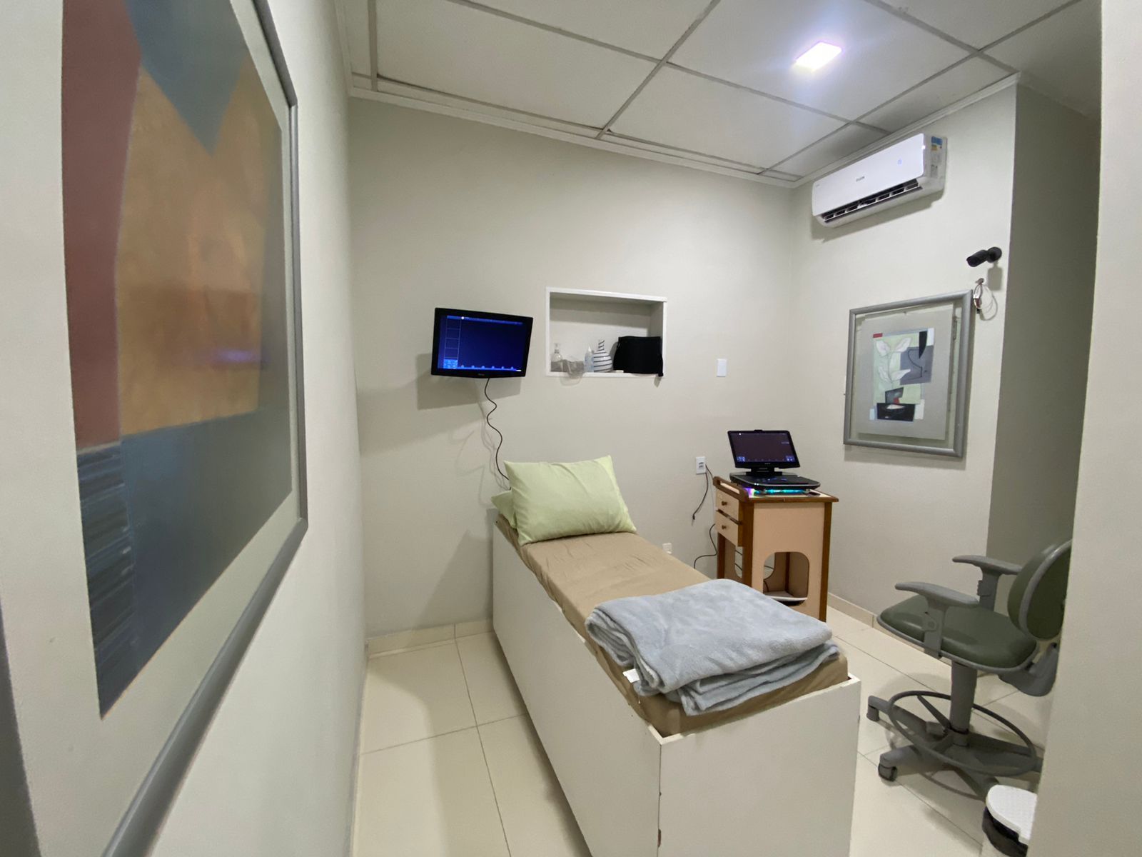 Sala de exames com cama, computador, obras de arte e ar condicionado.