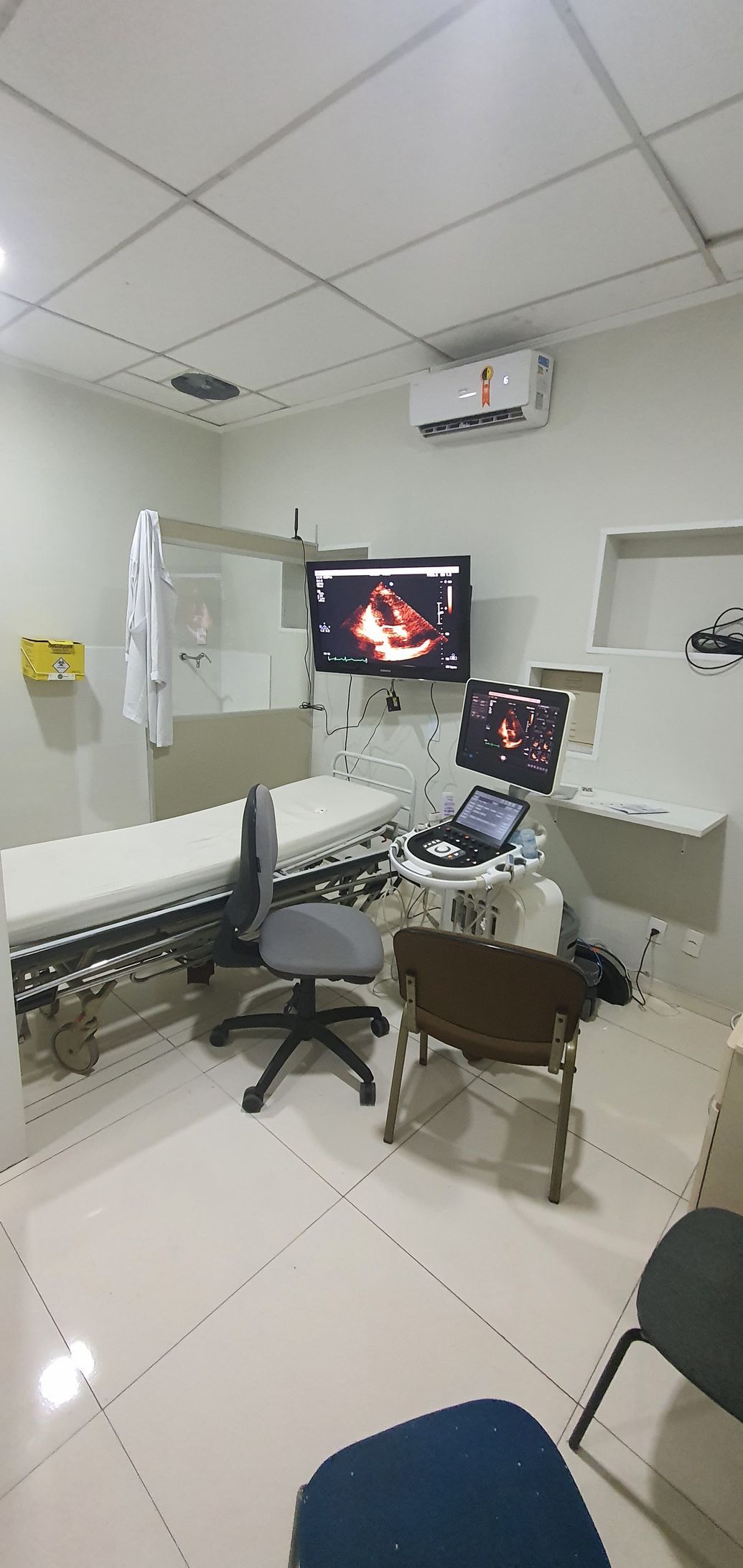 Sala de exames médicos com equipamento de ultrassom, maca e assentos.